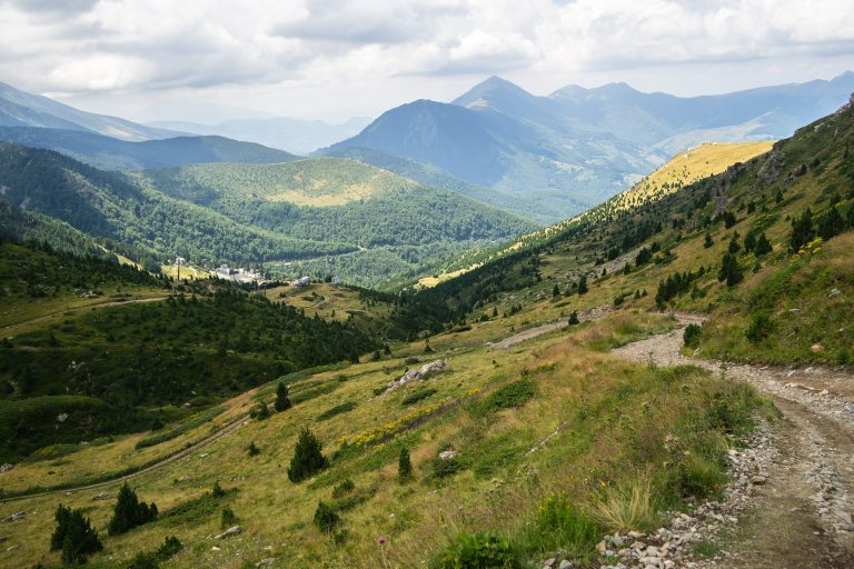 A landscape of hills covered in greenery with rocky mountains under a cloudy sky on the background